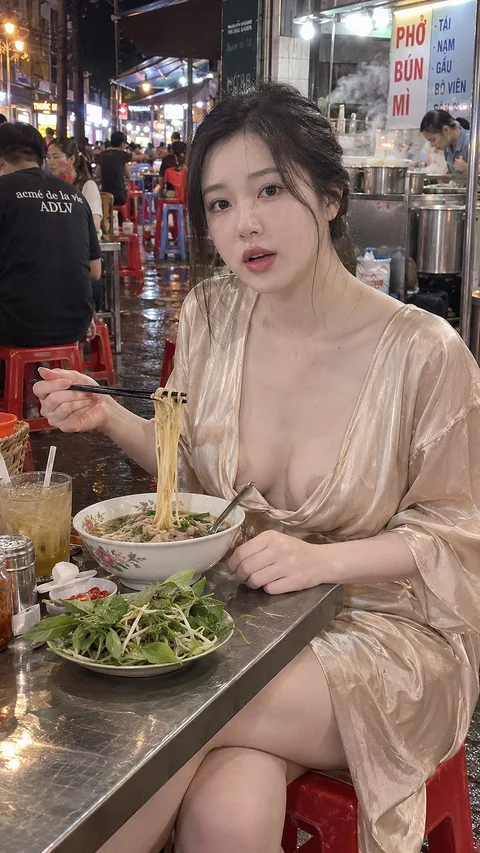A person in a pale satin outfit holds chopsticks above a bowl of noodles at a crowded food counter.
